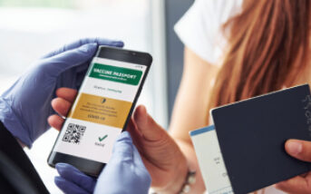 A close-up of a person holding a smartphone displaying a digital vaccine passport, while another person holds a physical passport and ticket. The person holding the smartphone is wearing blue gloves.