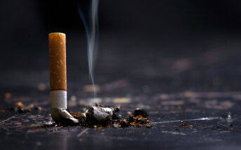 A cigarette butt is shown extinguished on a dark surface, with a small wisp of smoke still rising from it.